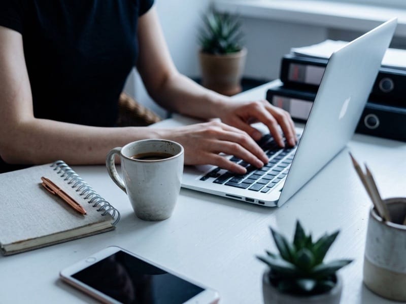 Woman typing on a laptop at a white desk with coffee mug, notebook, phone and succulents