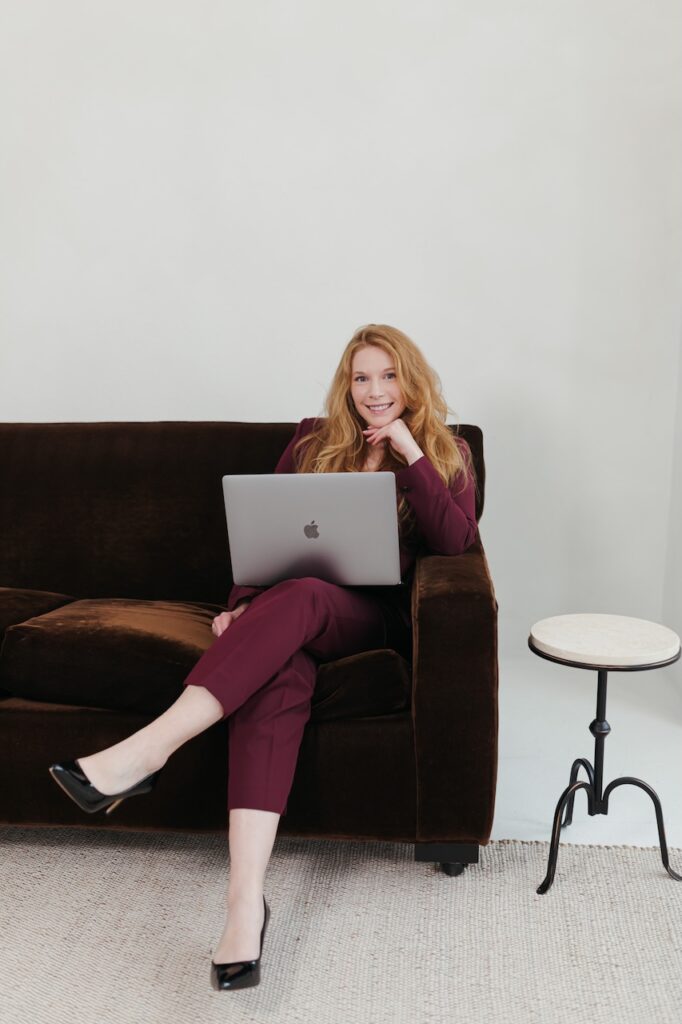 Cheryl Ann Barnes seated on a dark velvet sofa with an open MacBook, smiling at camera