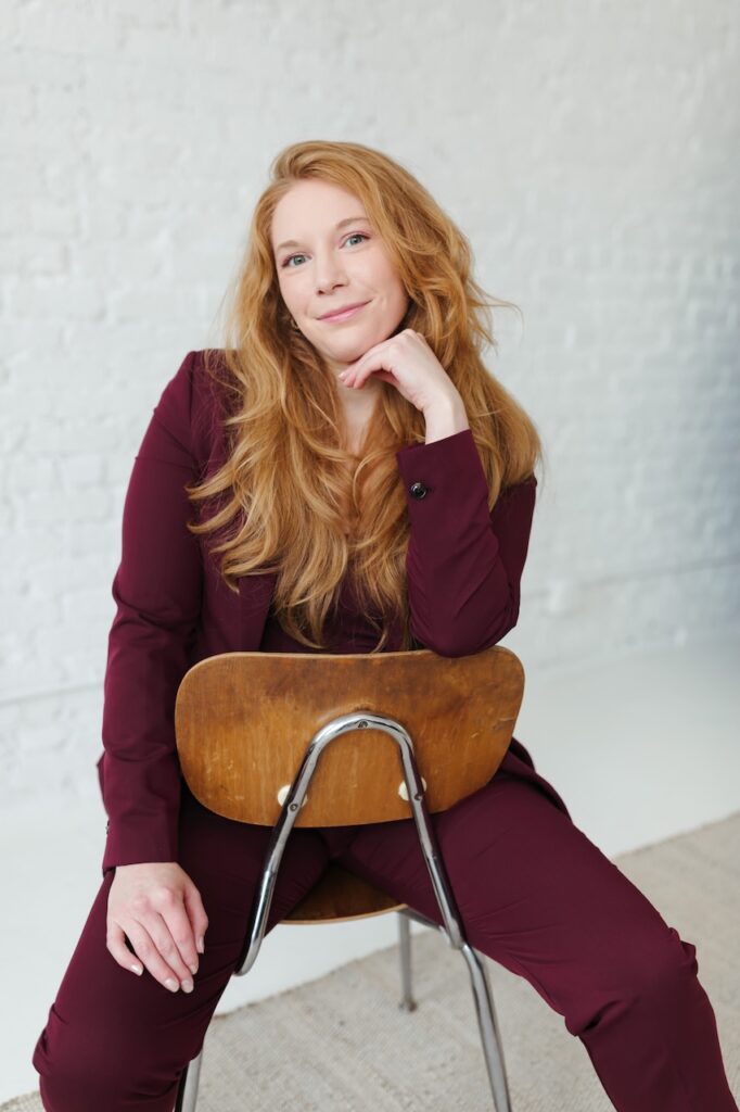 Cheryl Ann Barnes in a burgundy suit resting her chin on her hand, seated backward on a wooden chair