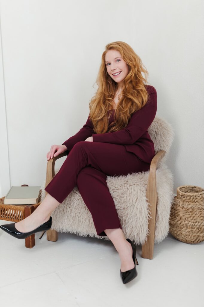 Cheryl Ann Barnes in a burgundy suit seated in a sheepskin armchair, smiling, with books and a wicker basket nearby