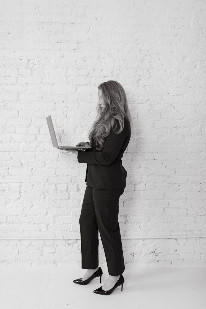 Cheryl Ann Barnes in a black suit standing in profile against a white brick wall, looking down at a laptop she holds open