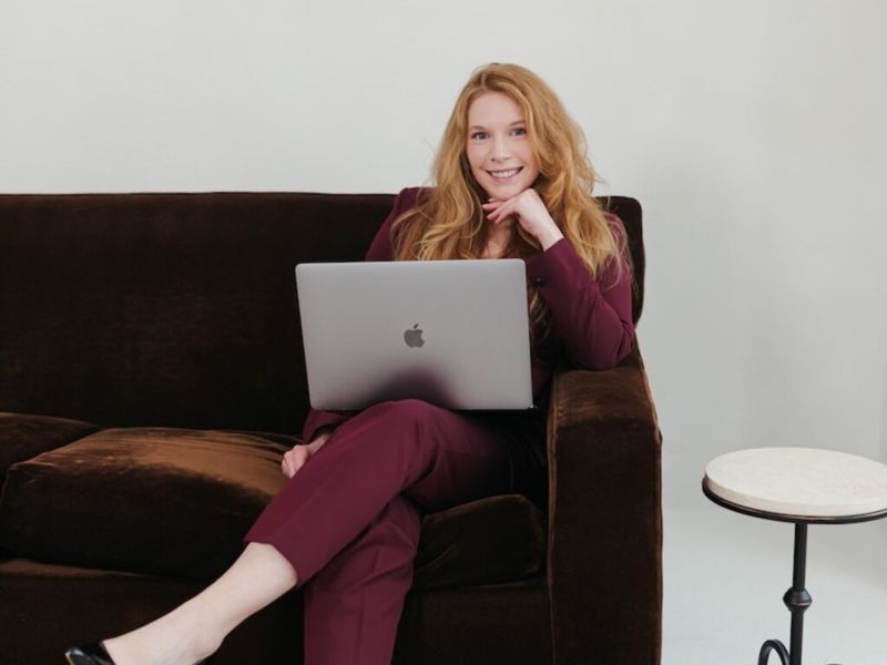 Cheryl Ann seated on a dark brown velvet sofa with a MacBook laptop, smiling at camera, wearing a burgundy suit