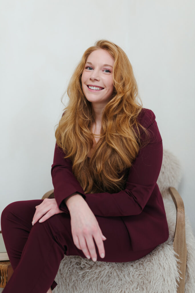 Cheryl Ann seated in a rattan chair wearing a burgundy suit, long auburn hair, smiling against a white studio background