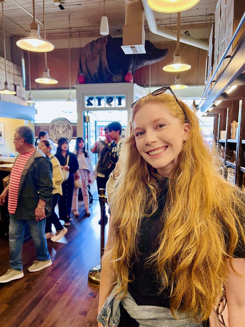 Woman with long red hair smiling inside a busy coffee shop with vintage Starbucks logo visible on the wall, pendant lights hanging from industrial ceiling