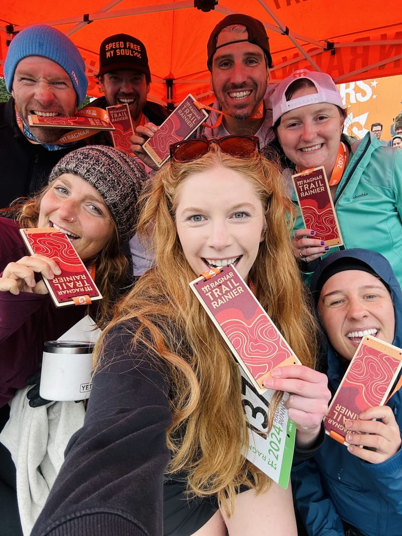 Group selfie of eight smiling runners holding up Ragnar Trail Rainier finisher medals under an orange tent, faces packed together in celebration