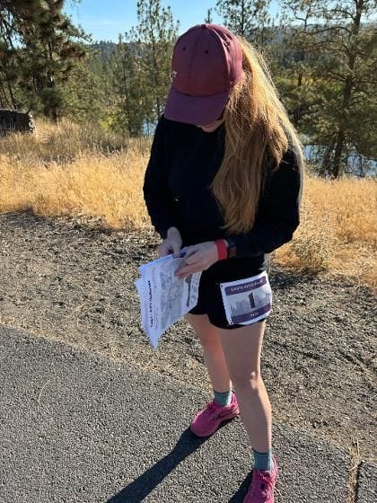 Runner stopped on trail looking down at a race course map, wearing black long sleeve and pink cap with dry grass and evergreen trees behind