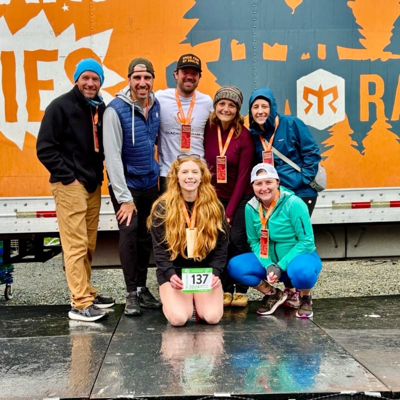 Group of seven runners posing together in front of an orange Ragnar relay trailer on wet pavement, wearing medals and race gear in cool weather