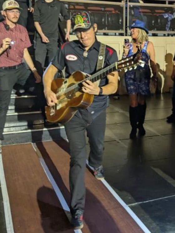Stephen Wilson Jr. walking through the crowd playing acoustic guitar, wearing a dark work shirt and colorful cap, with fans watching nearby